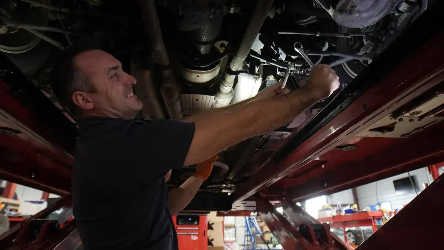 Mechanic working under a car on a lift; holding a tool, face strained with effort, in a garage.