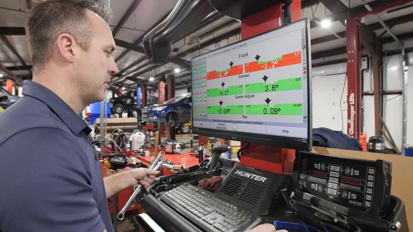 Mechanic adjusting a car's alignment using a computer display. Green and orange data appears on screen in a garage setting.