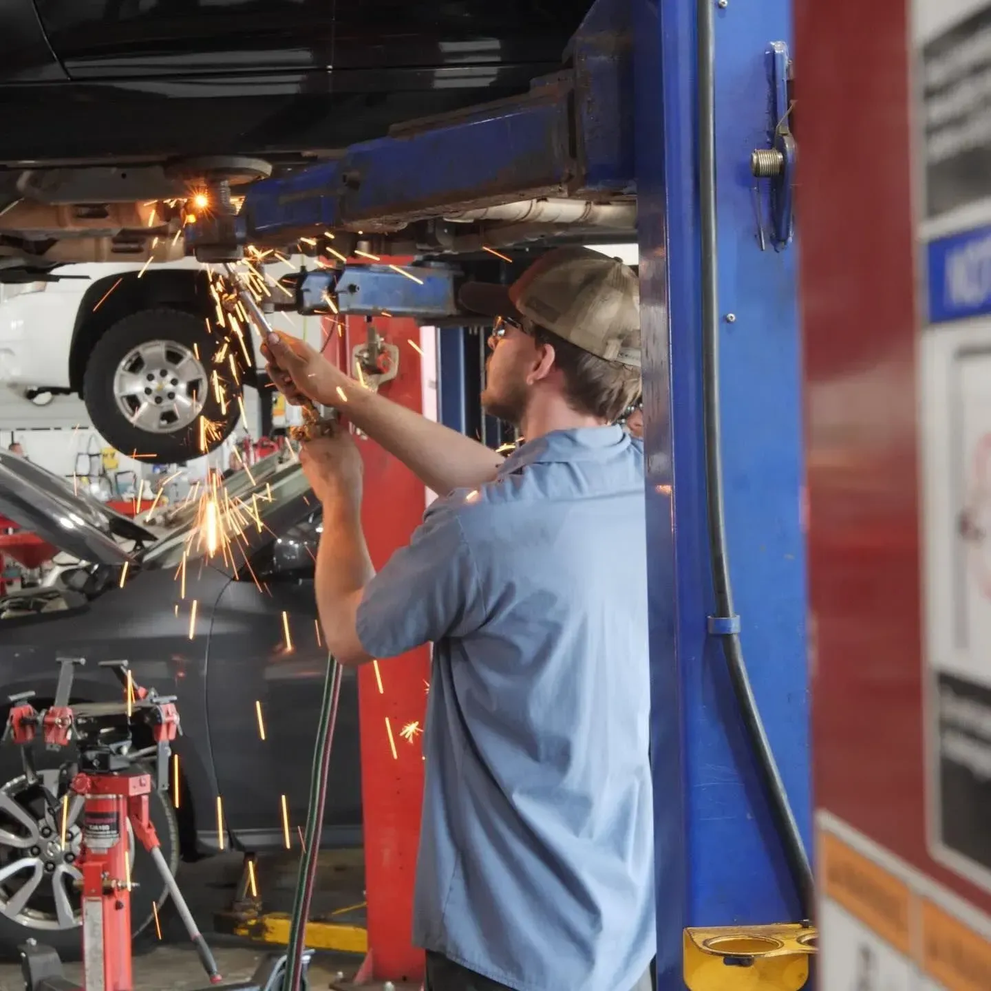 Mechanic working under a car on a lift; sparks flying. Shop interior, blue shirt, baseball cap.