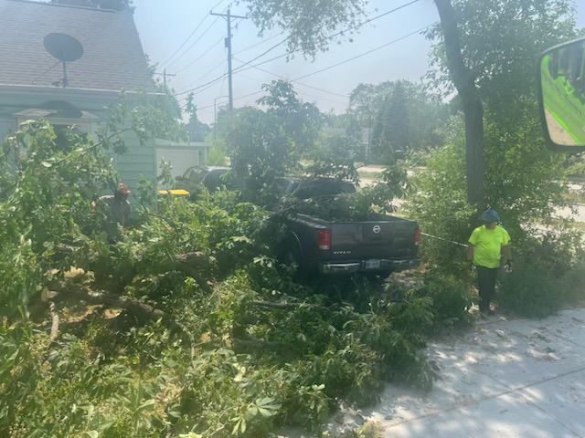 A truck is heavily damaged by fallen trees, likely from a storm. Two people wearing safety vests are working to clear the debris.