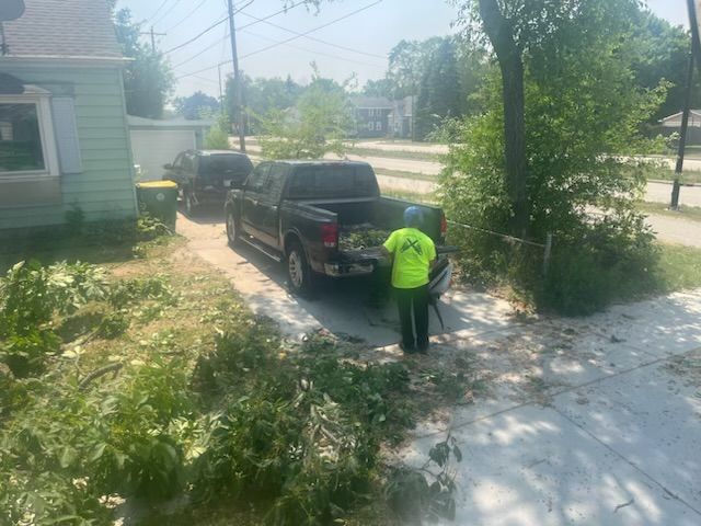 A worker loads brush into a pickup truck on a driveway. The worker wears a neon green shirt.