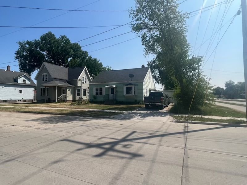 Two-story light green houses and a gray pickup truck sit on a dusty lot under a sunny sky. Power lines and tree shadow cross the road.