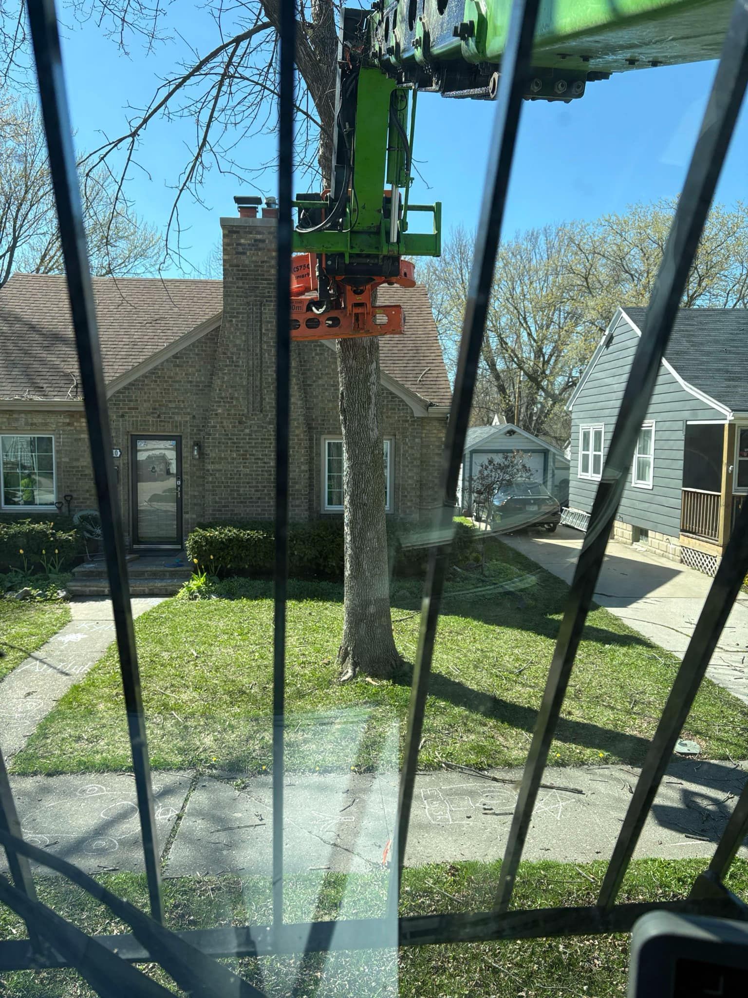 View from inside a tree trimming machine; machine's cutting arm is positioned to cut a tree in front of a house on a sunny day.