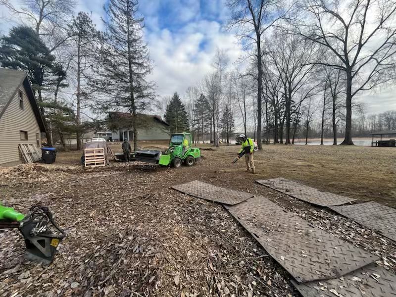 A person uses a leaf blower near a green machine on a lawn.  Brown leaves cover the ground, and trees line the perimeter.