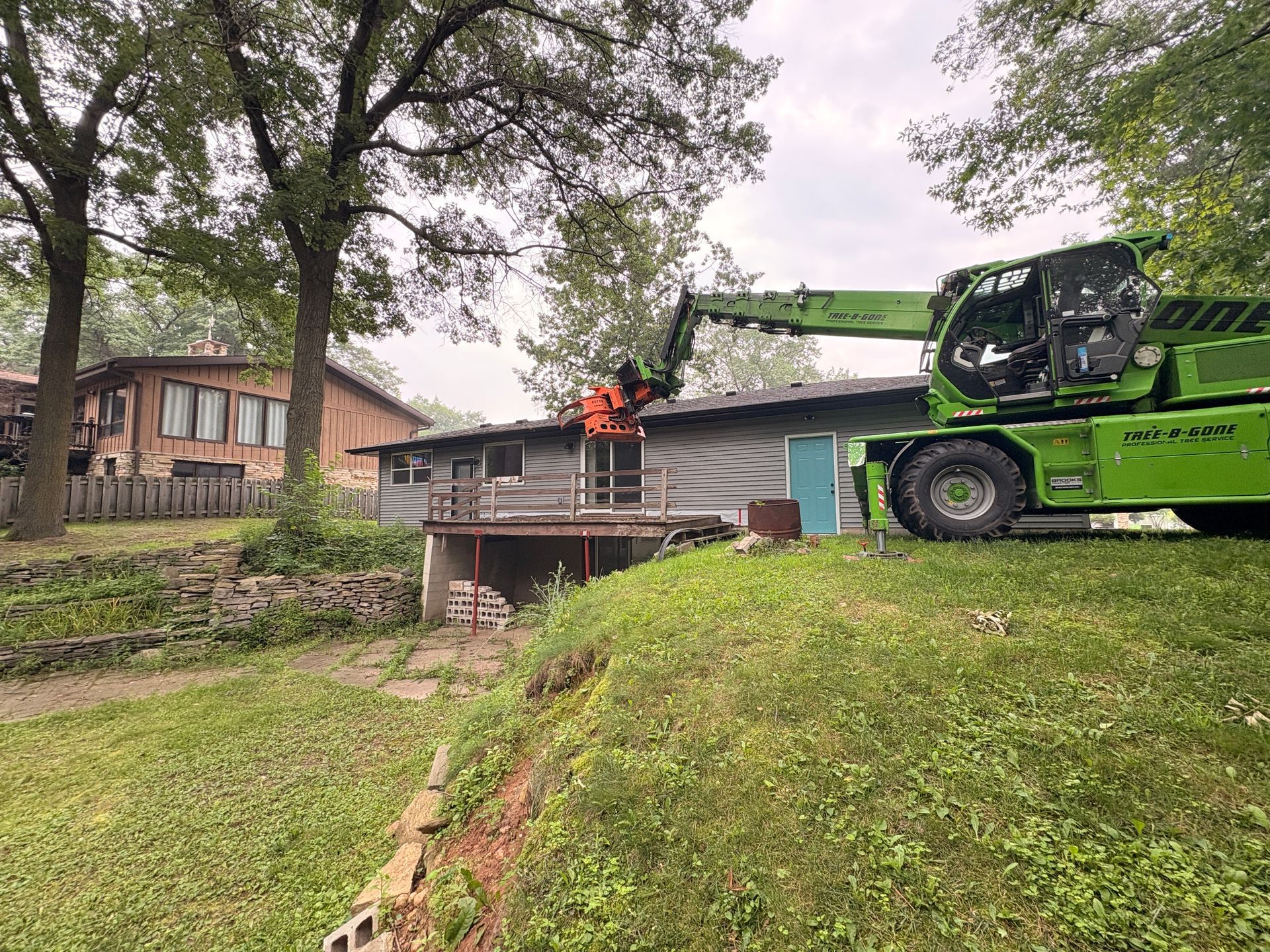 Green telehandler dismantling a small, dilapidated building near a grassy hill and residential homes.
