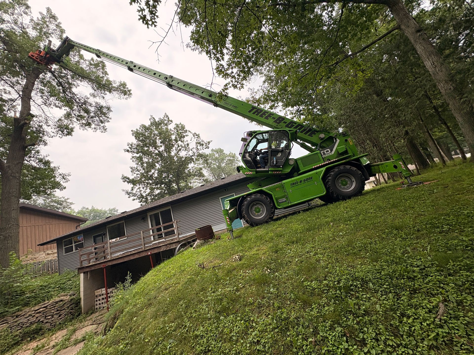 Green tree-trimming machine cutting branches from a tree next to a house on a hillside.