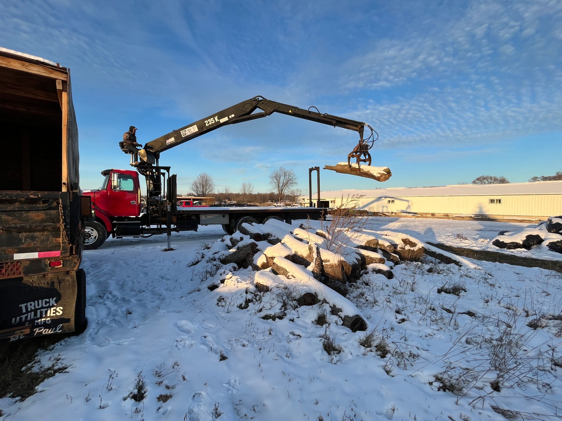 A red truck with a crane lifts a wooden beam on a snowy field. A person is visible operating the crane.