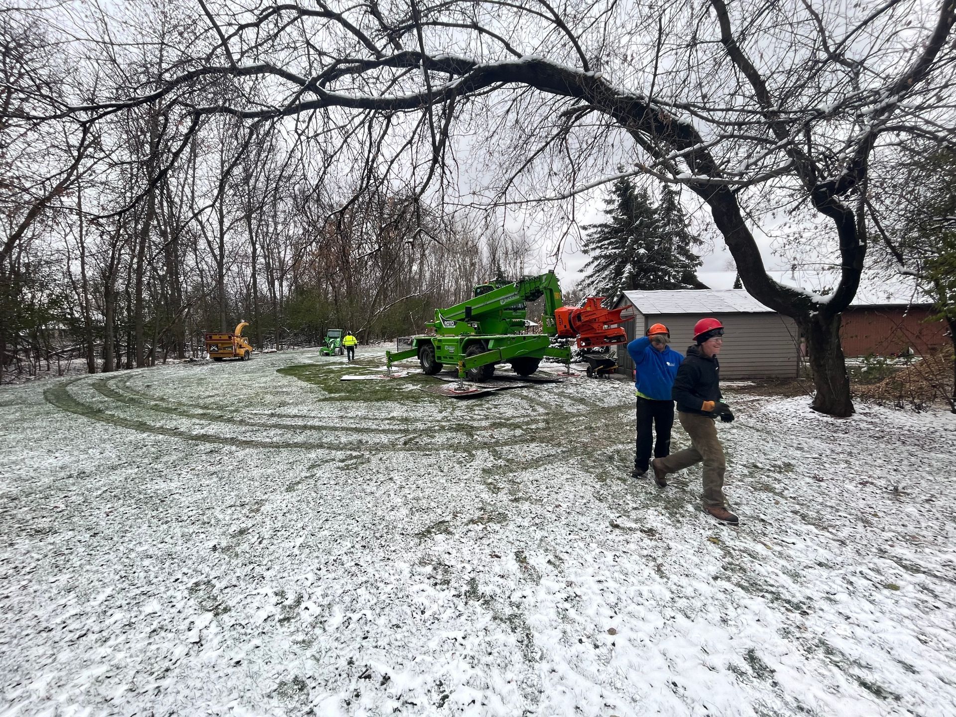 Snowy scene with tree removal equipment, including a green lift and orange chipper. Workers in hard hats are present.