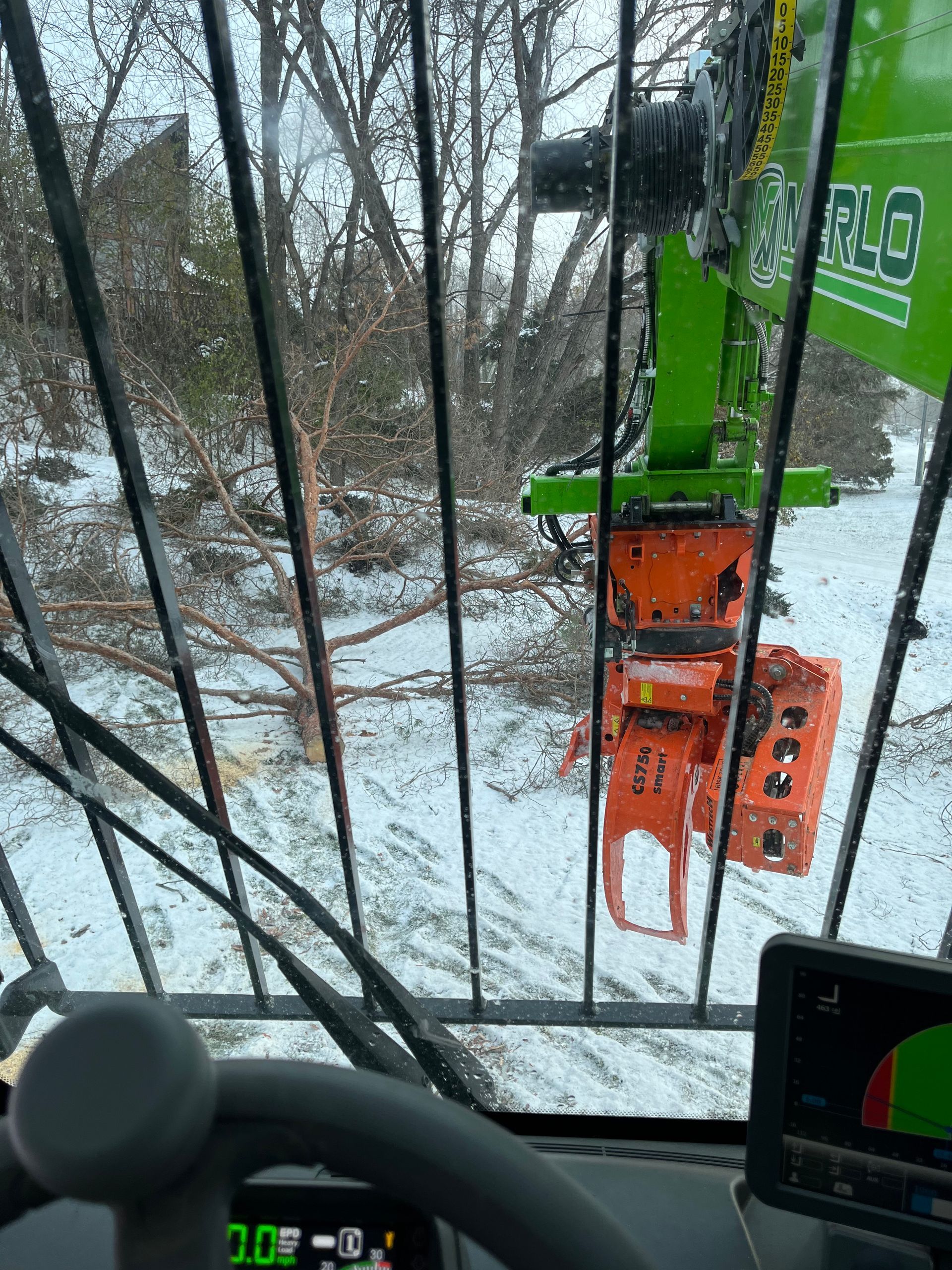 View from inside a logging machine's cab; a green arm extends toward a tree in a snowy forest.