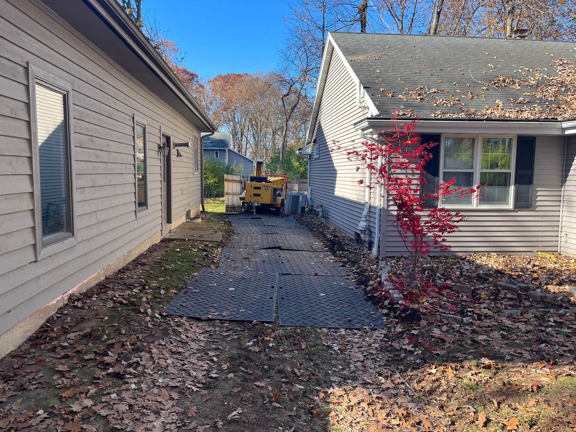 A narrow backyard path between two houses with black grids on the ground, covered in fallen leaves. A tree grinder is in the distance.