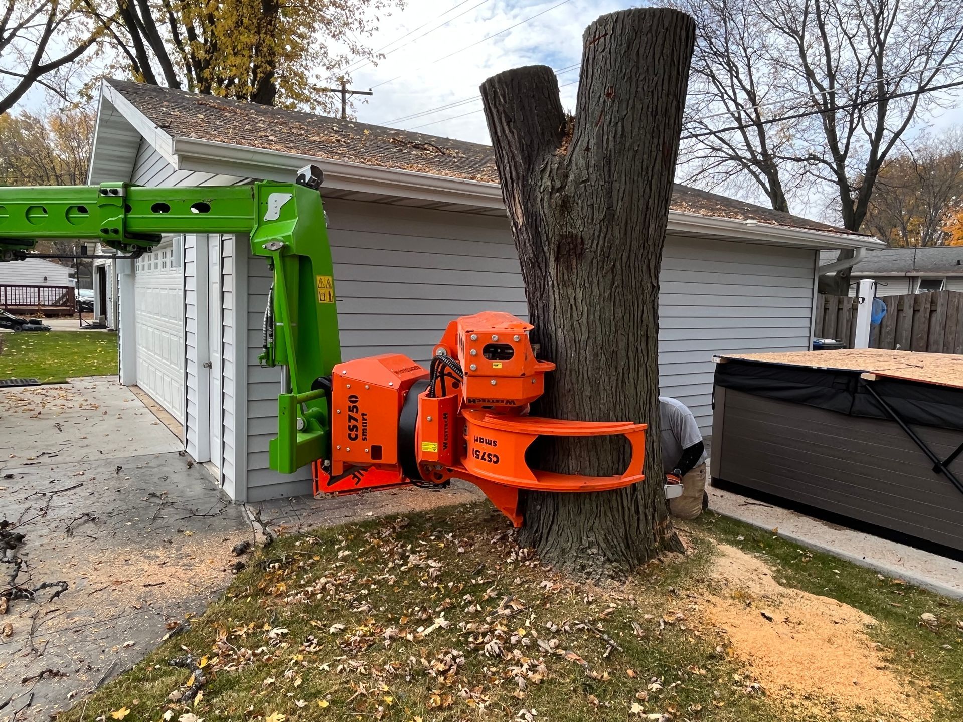 A tree is being cut with a tree-cutting machine attached to a green crane next to a white garage.