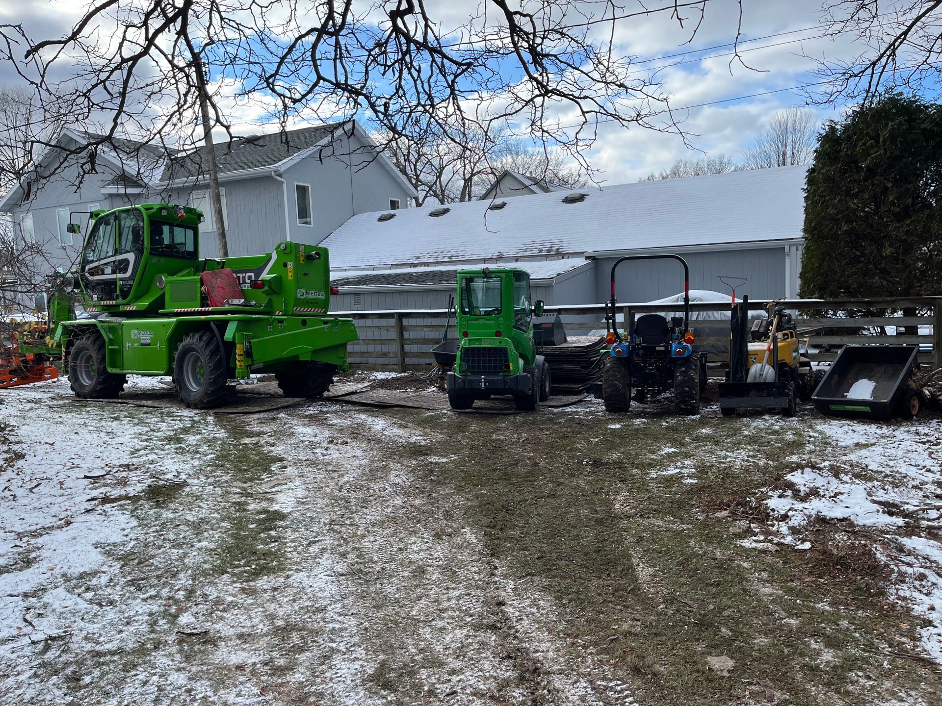 Several green construction vehicles and other machinery sit on a snowy, muddy yard near a house on a winter day.