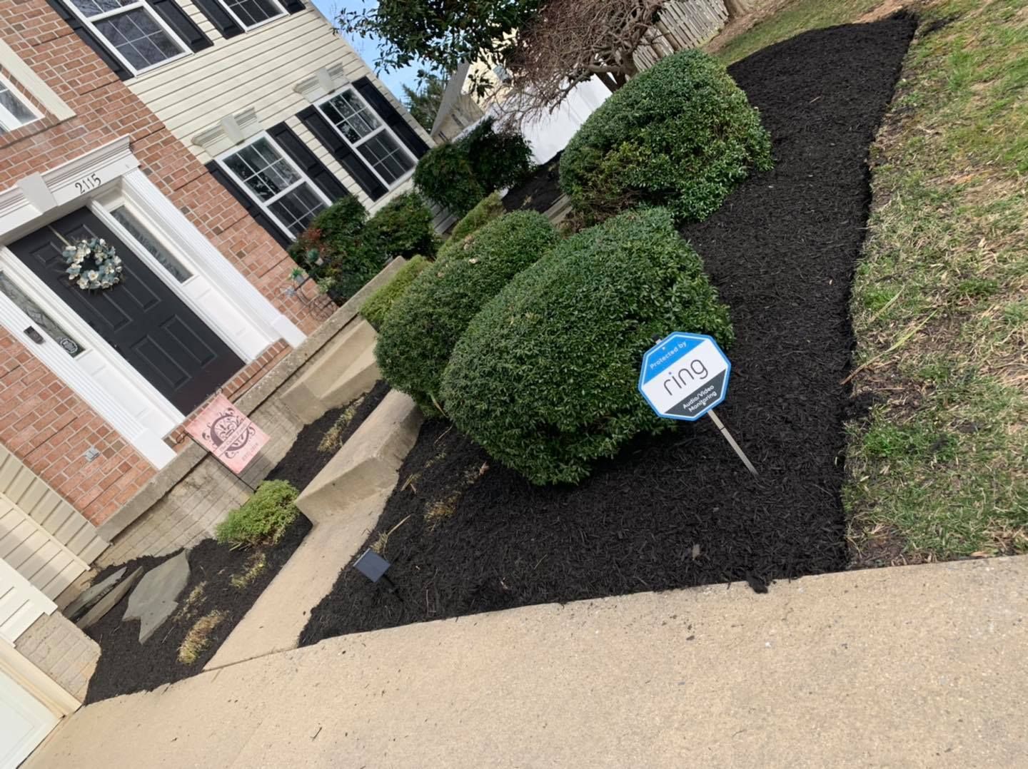 Front yard with black mulch, neatly trimmed shrubs, and a house entrance with a porch sign.