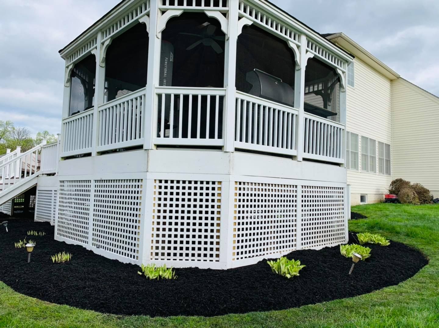 White screened porch with lattice skirting beside a beige house, bordered by black mulch and small shrubs