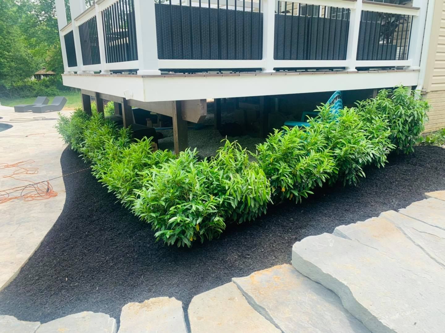 Raised porch corner with white railing, black skirting, and green shrubs beside cracked concrete walkway