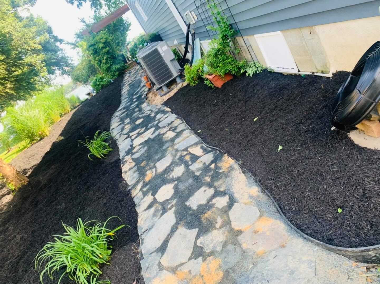 Stone walkway beside a house, bordered by black mulch, small plants, and an air conditioner unit.