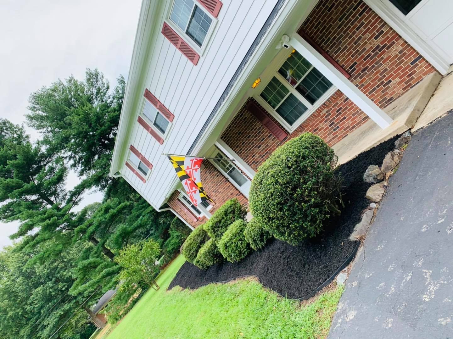 Brick house entrance with red shutters, colorful hanging sign, and trimmed bushes beside a driveway.