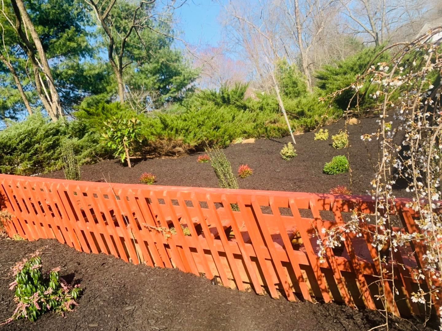Orange fence along a mulched garden hillside with shrubs and trees under a blue sky