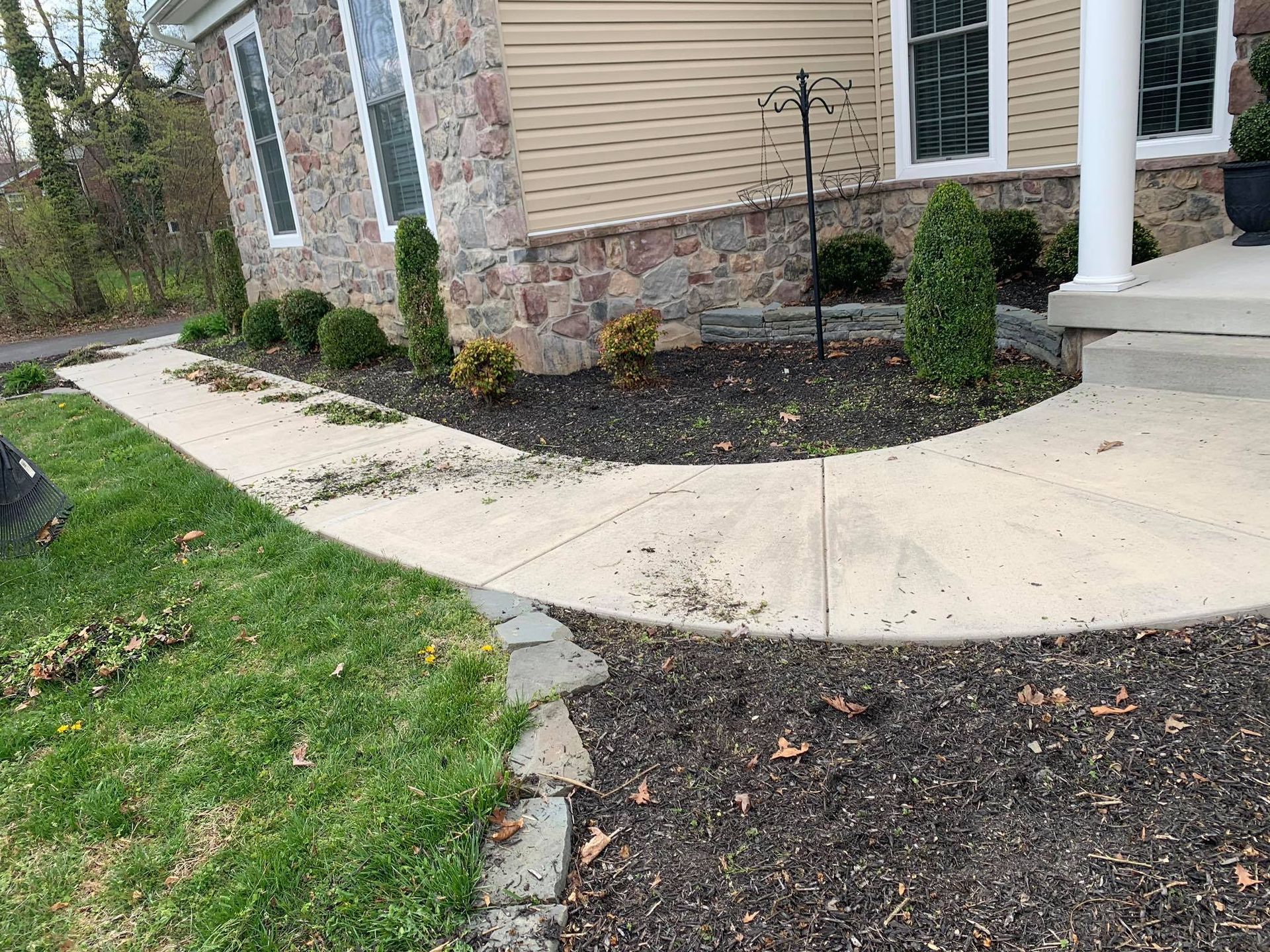 Curved concrete walkway beside a beige house with stone trim and mulch beds