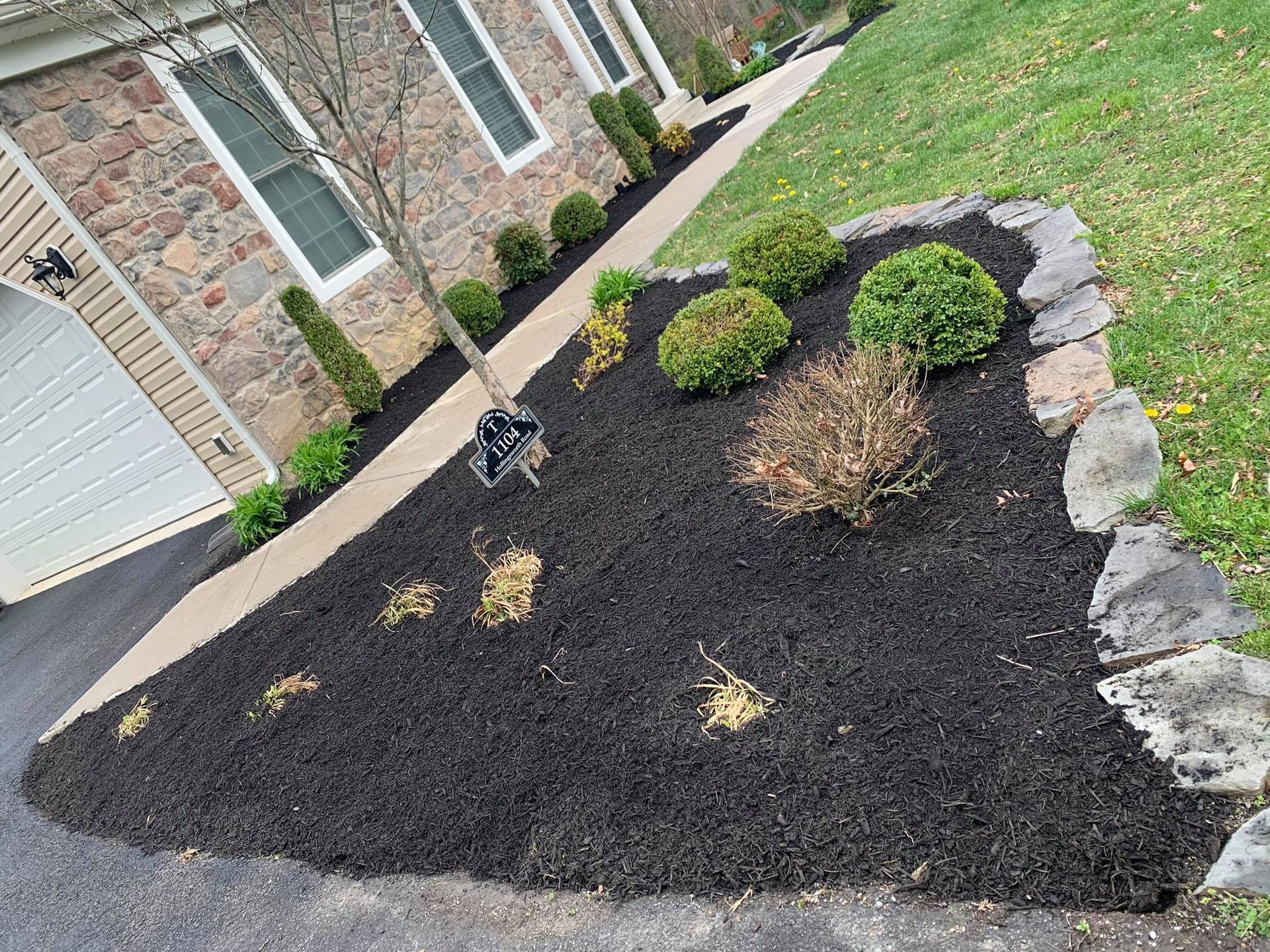Front yard landscaped bed with black mulch, small shrubs, and stone edging beside a house.