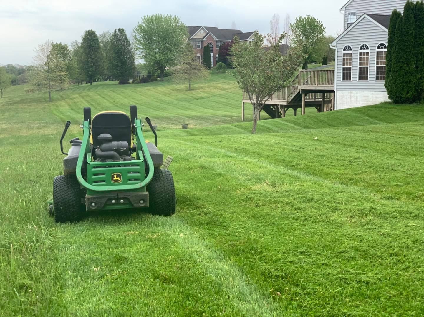 Green riding lawn mower on a grassy yard near a white house and trees.