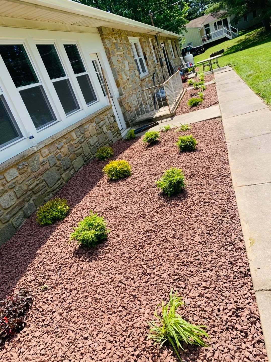 Landscaped side yard with red mulch, small shrubs, stone house wall, and concrete walkway under bright sun