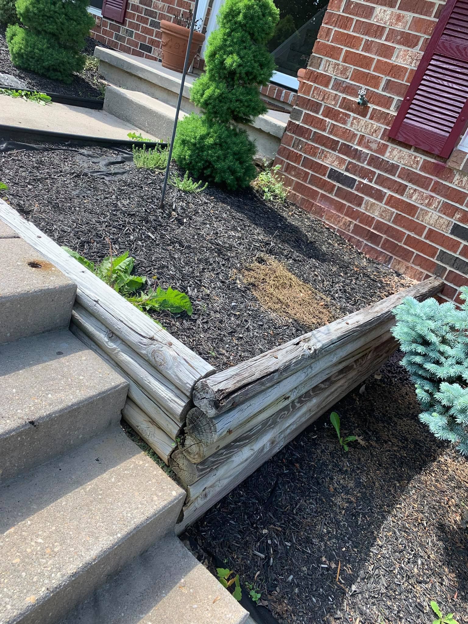 Wooden retaining wall beside concrete steps, with gravel beds and brick house exterior.