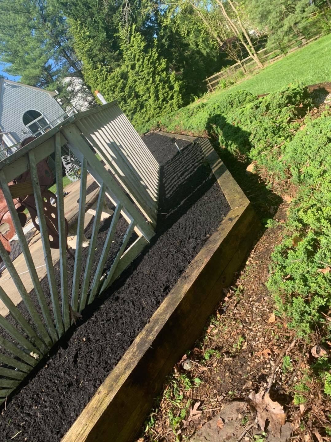 Sloped backyard with a raised deck, brick retaining wall, and green bushes in sunlight.