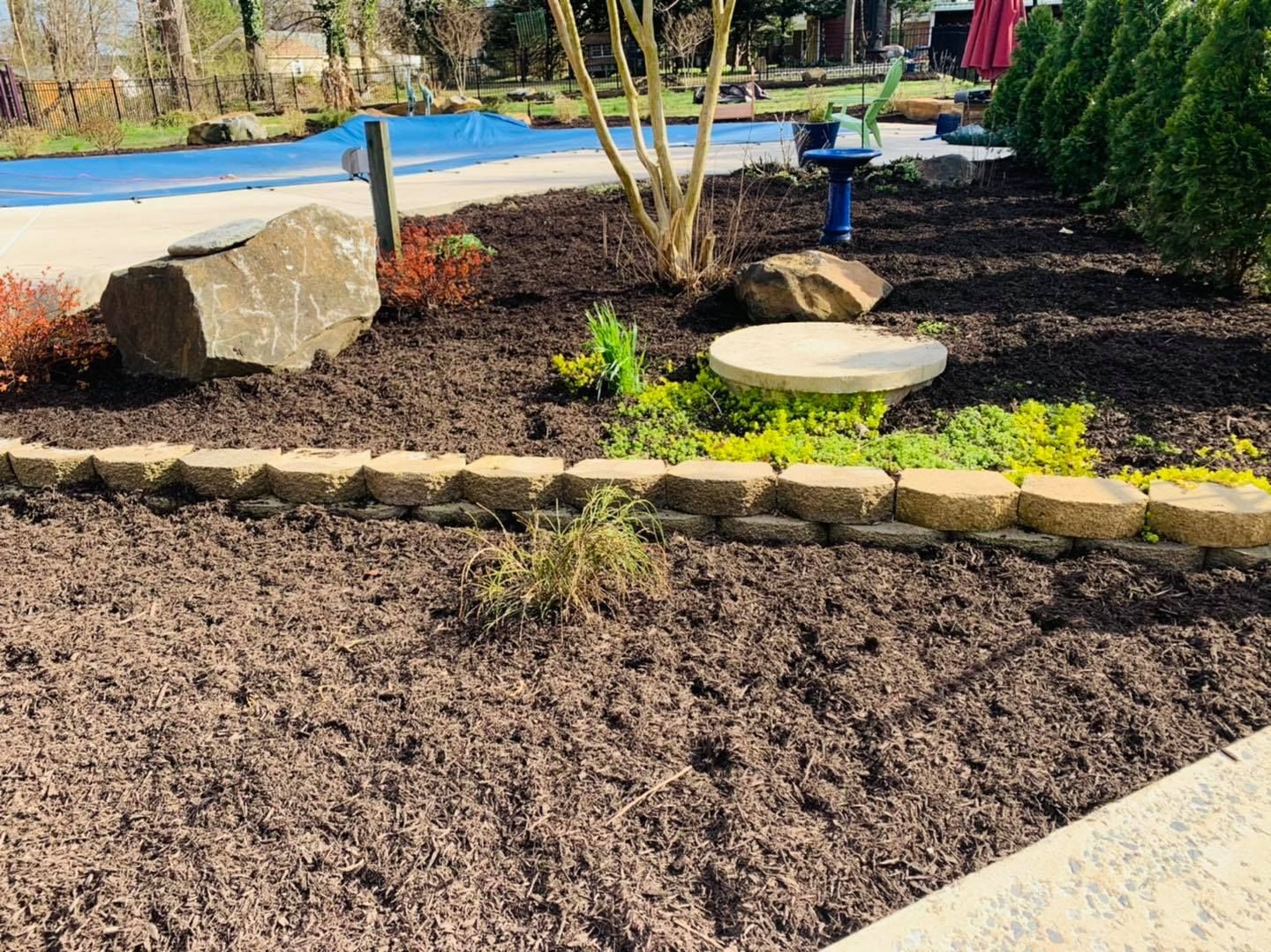 Landscaped yard with mulch beds, stone edging, rocks, and a pool in the background.