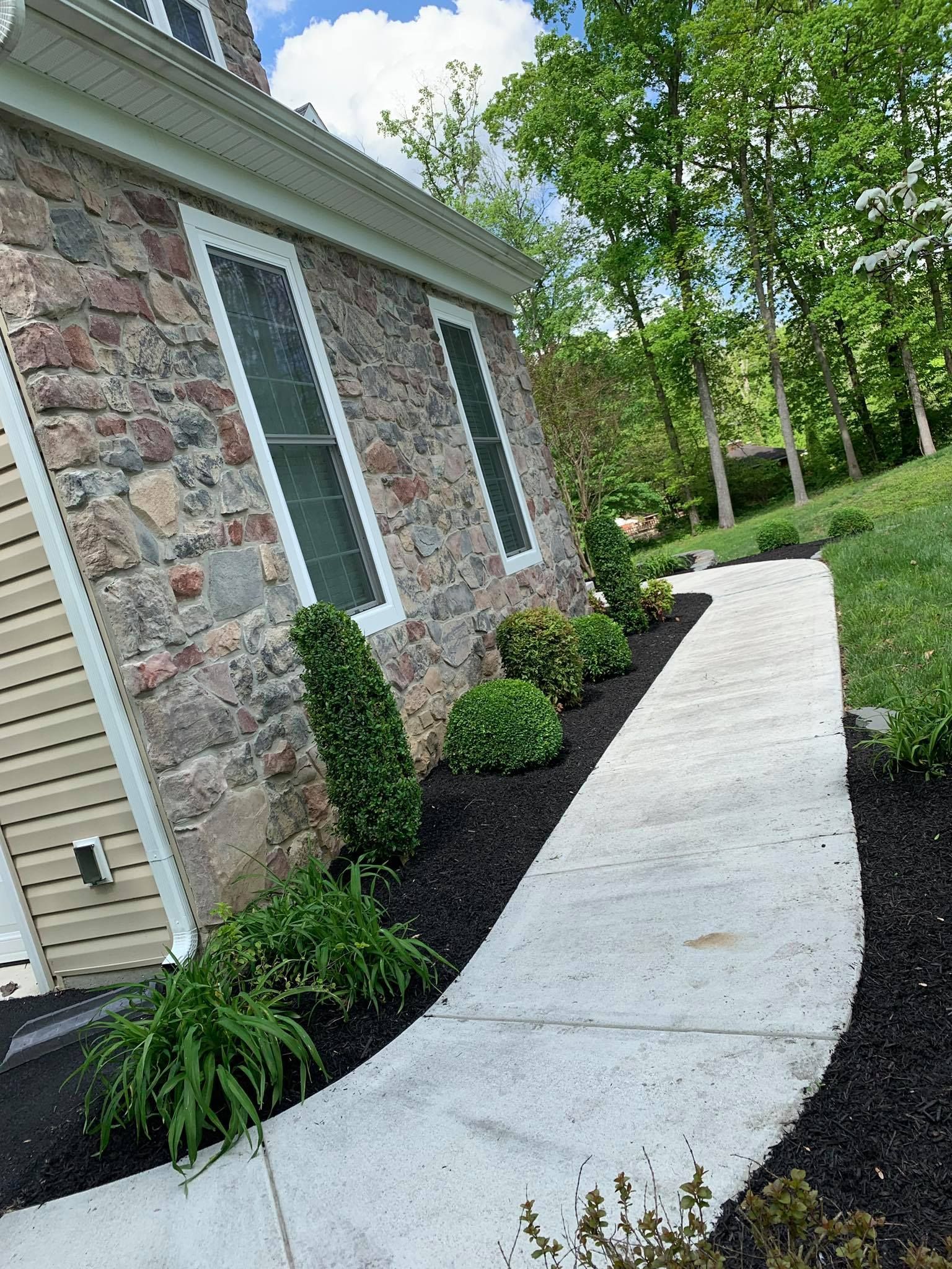 Curved concrete walkway beside a stone house with windows and landscaped shrubs