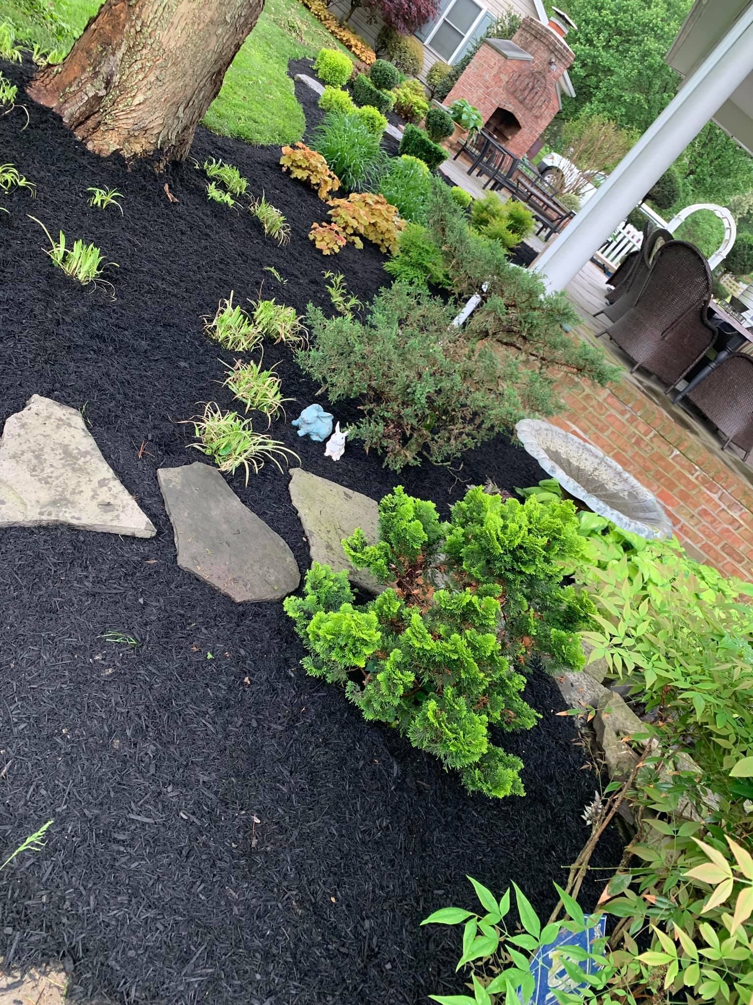 Mulched garden bed with shrubs, stepping stones, and a brick house in the background