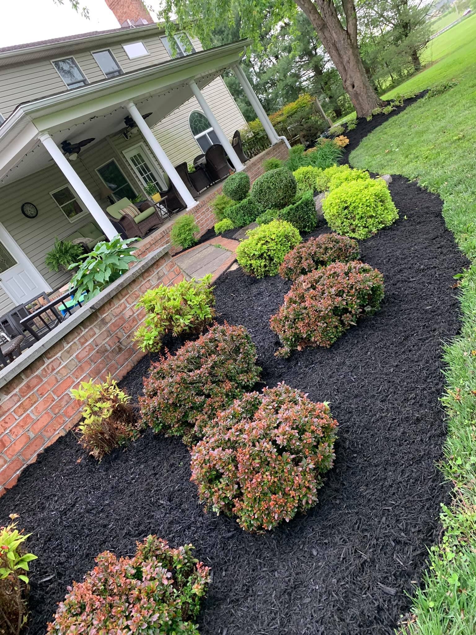 Front yard garden with black mulch, trimmed shrubs, brick edging, and a house porch in the background