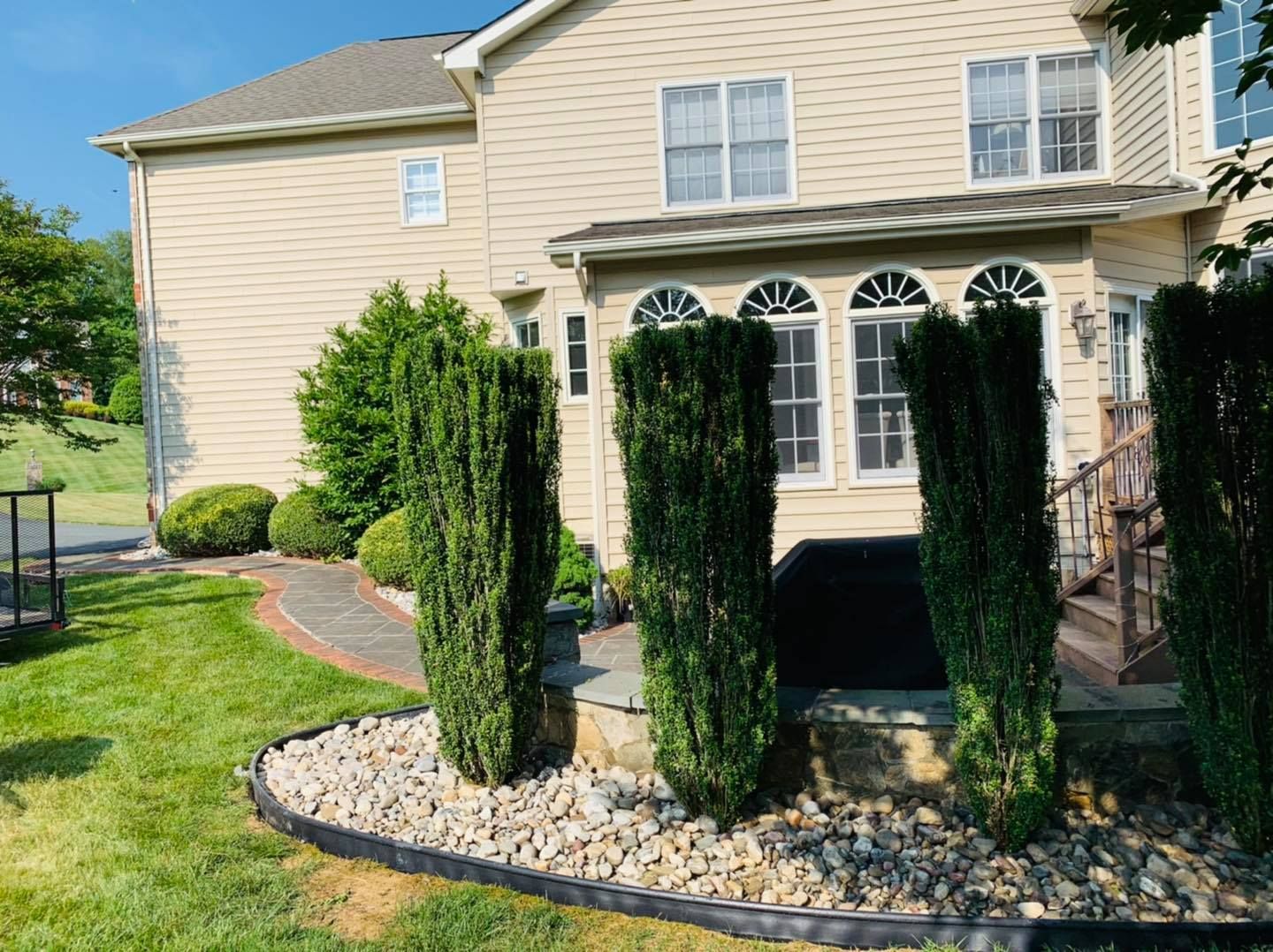 Backyard patio with beige house, arched windows, and two tall evergreen shrubs in a stone border