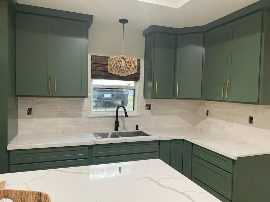 A kitchen with green cabinets , white counter tops , a sink and a window.