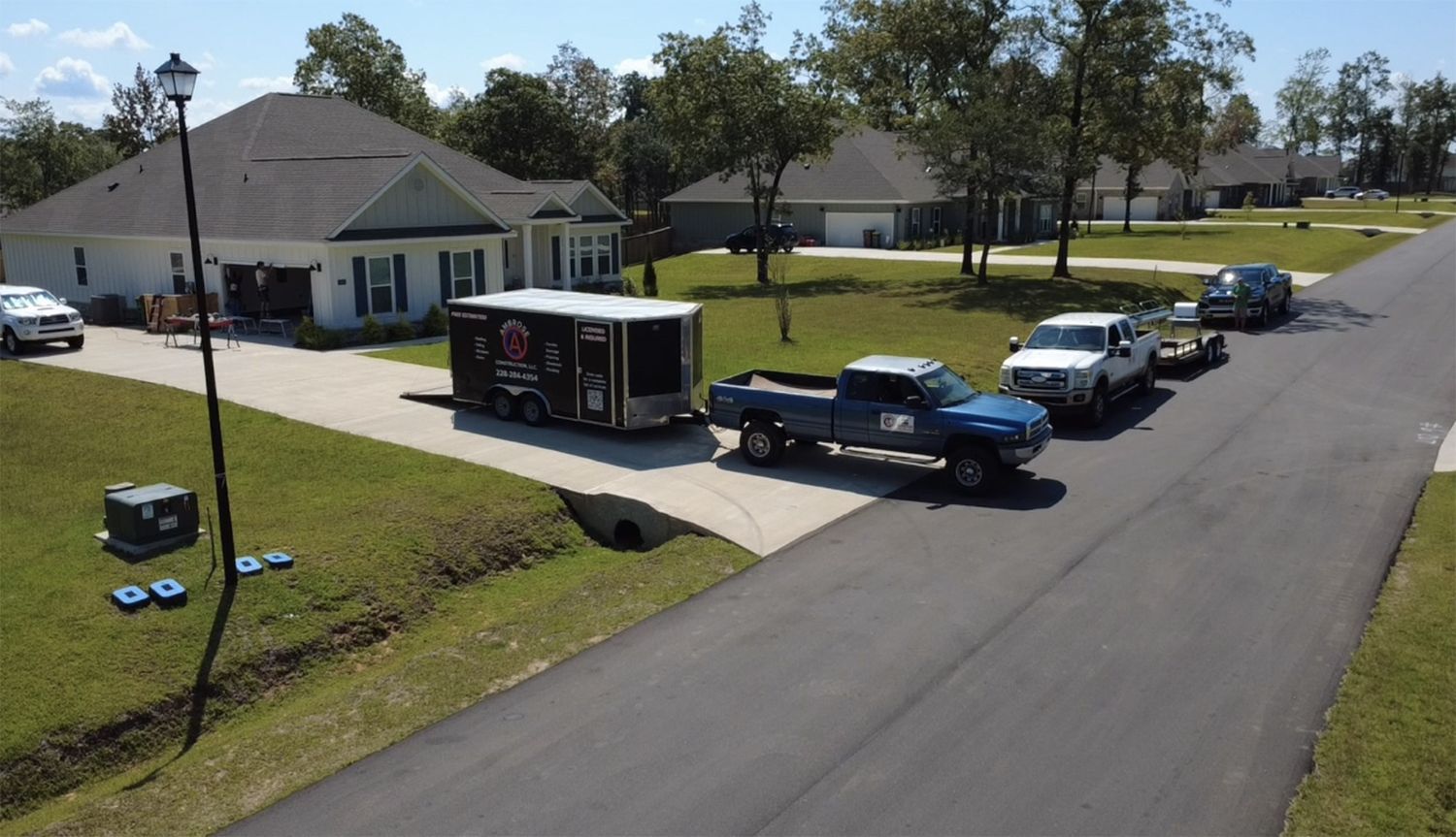 A blue truck pulling a trailer in a suburban street, with a white house in the background and other vehicles.