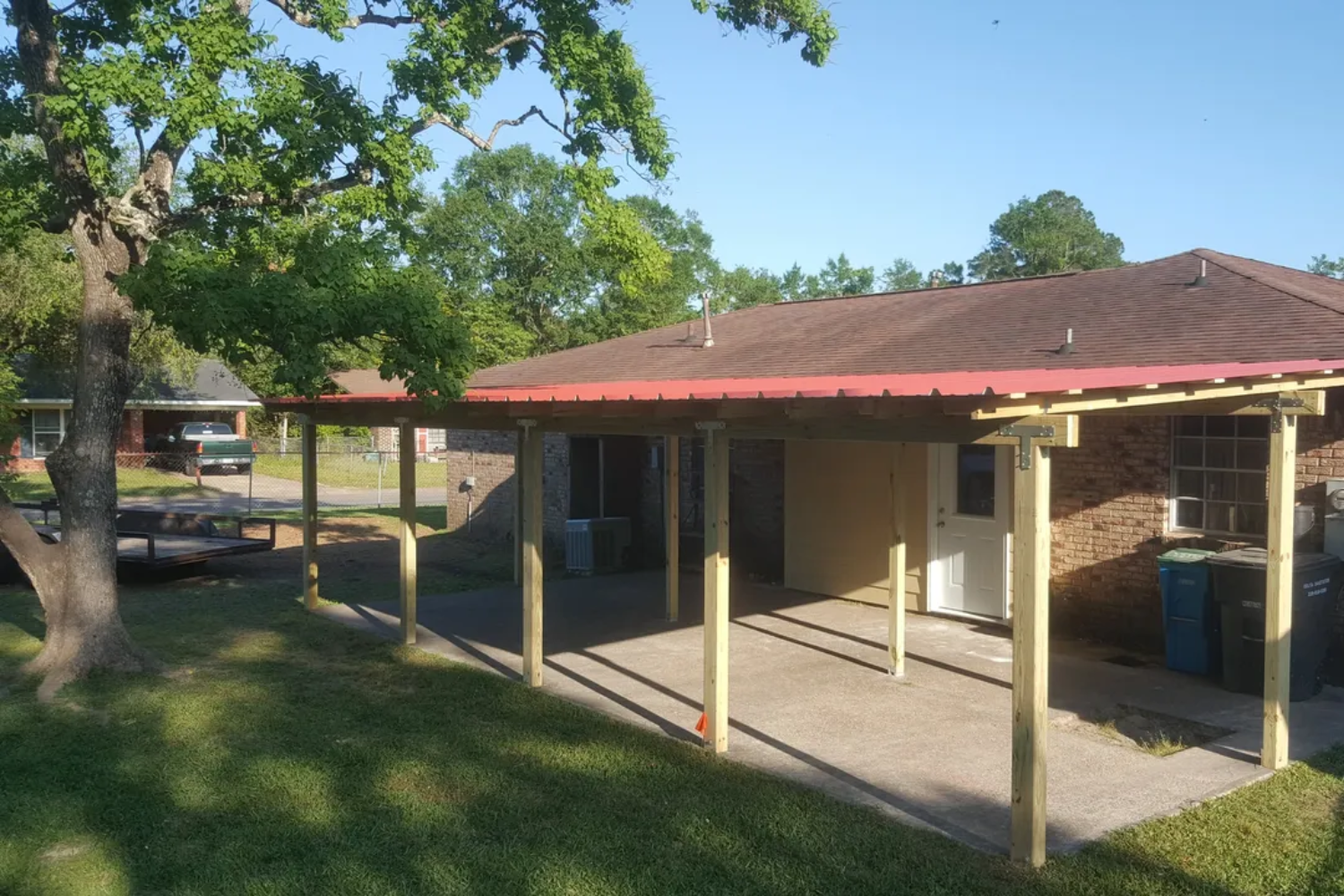 A covered patio with red roof, supported by wooden posts, extending from a brick house.
