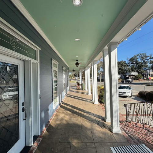 A porch with white pillars and a green ceiling. Gray building with windows and a view of the street.