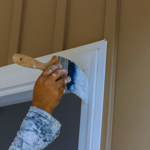 Hand painting white trim of a door frame with a brush. Beige siding in the background.