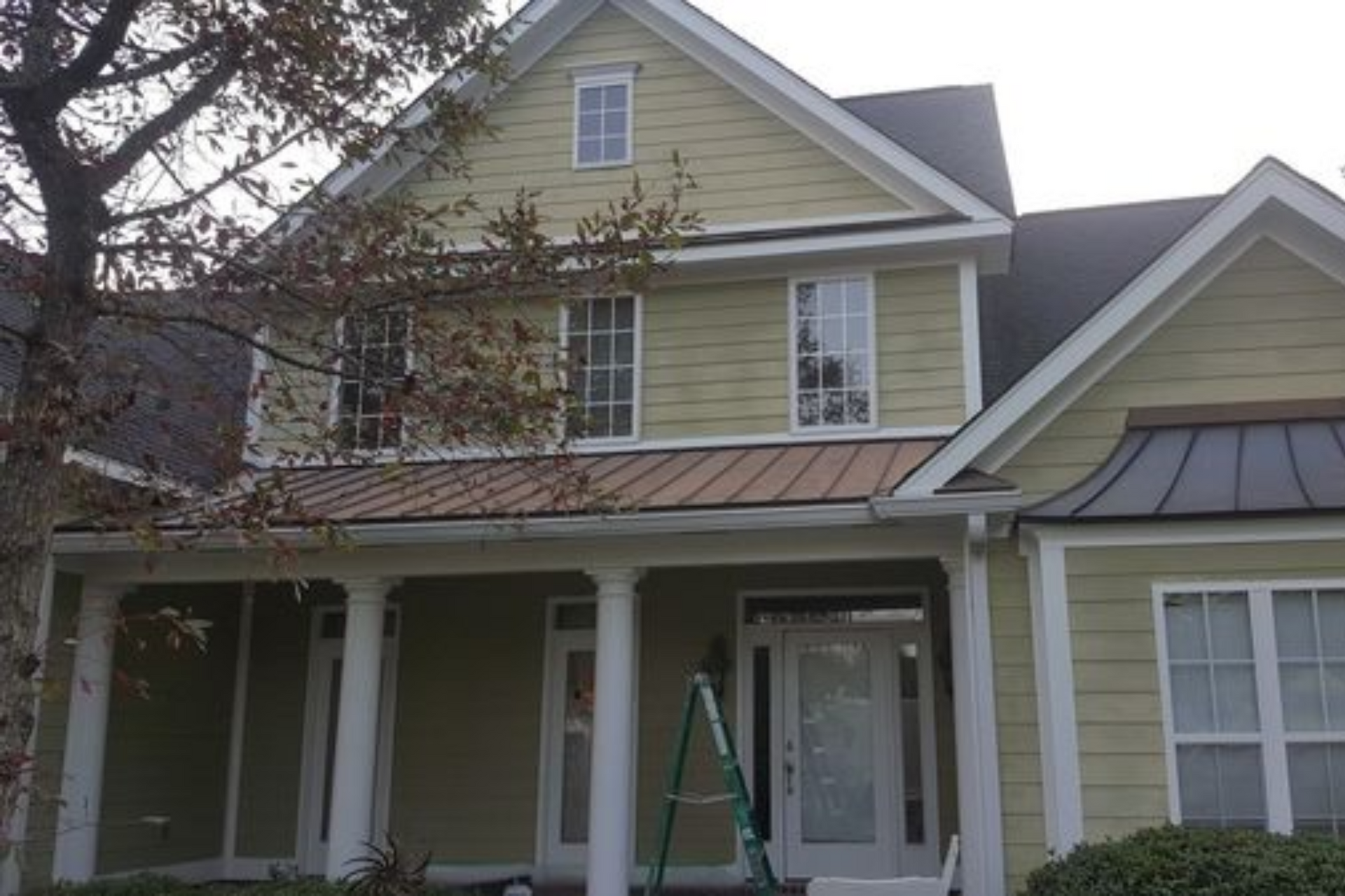 Two-story house with green siding, white trim, and a brown porch roof. A ladder leans against the door.