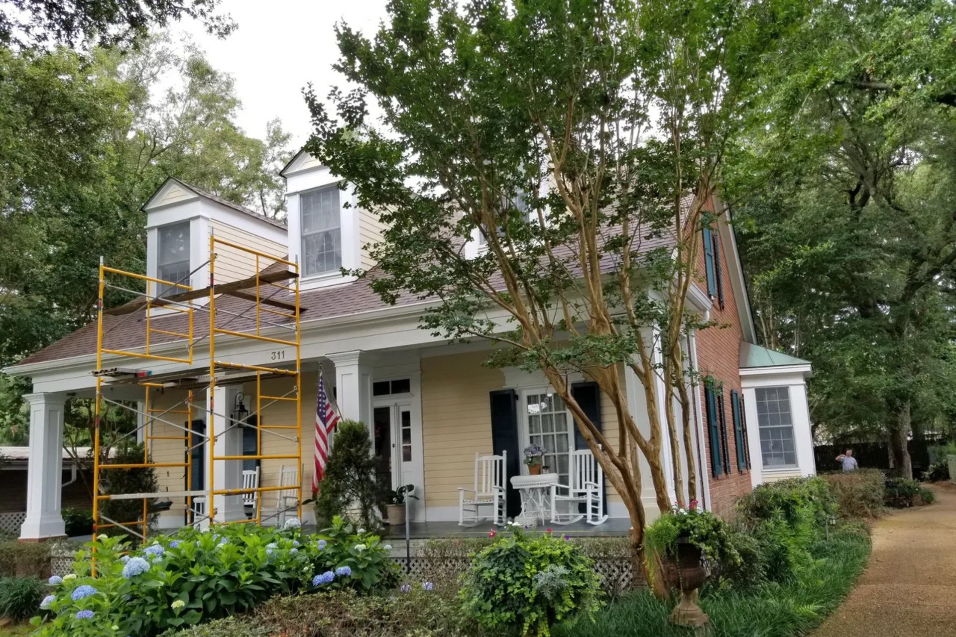 A two-story house with a porch, scaffolding, and a US flag. Exterior is tan and brick, surrounded by trees.