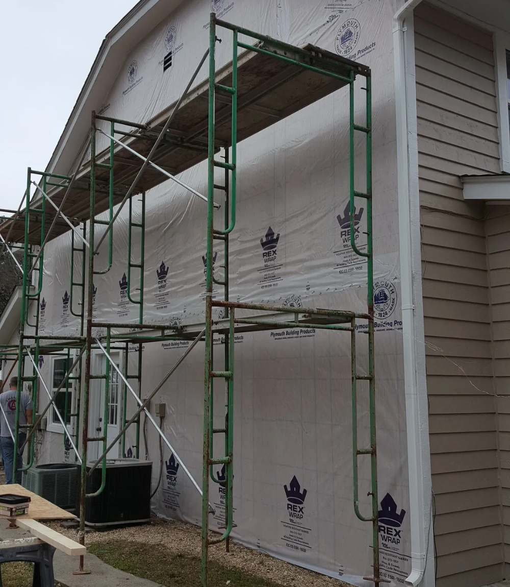 Scaffolding on house with green frame, white wrap, and tan siding.