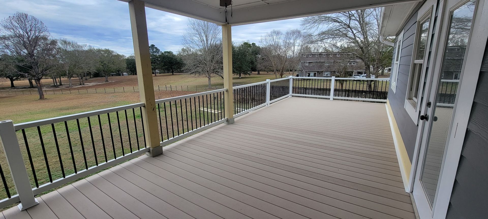 A covered porch with a wooden deck overlooking a grassy field and trees on a cloudy day.