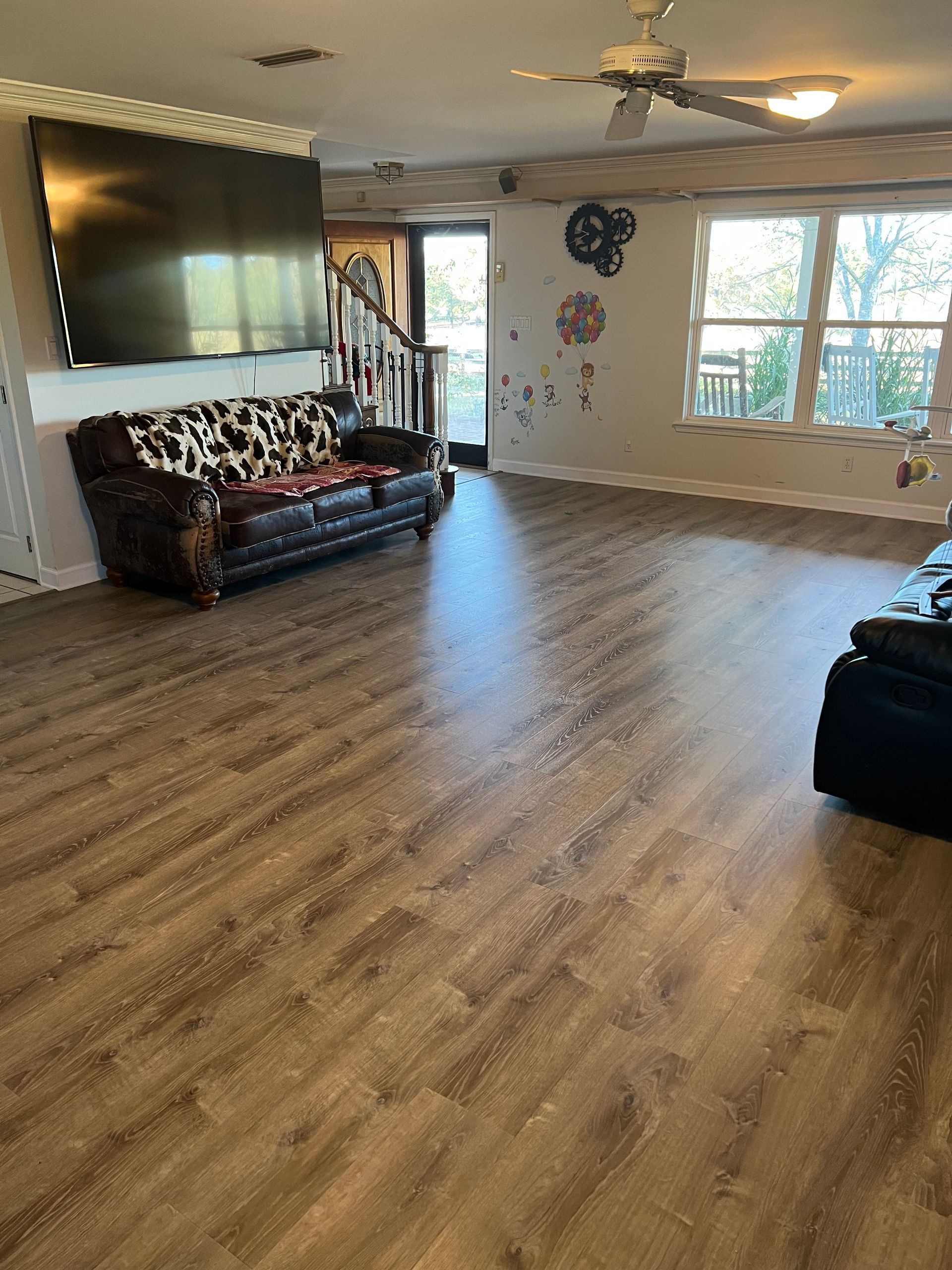 Living room with brown wood-look flooring, cow-print couch, large TV, and windows.