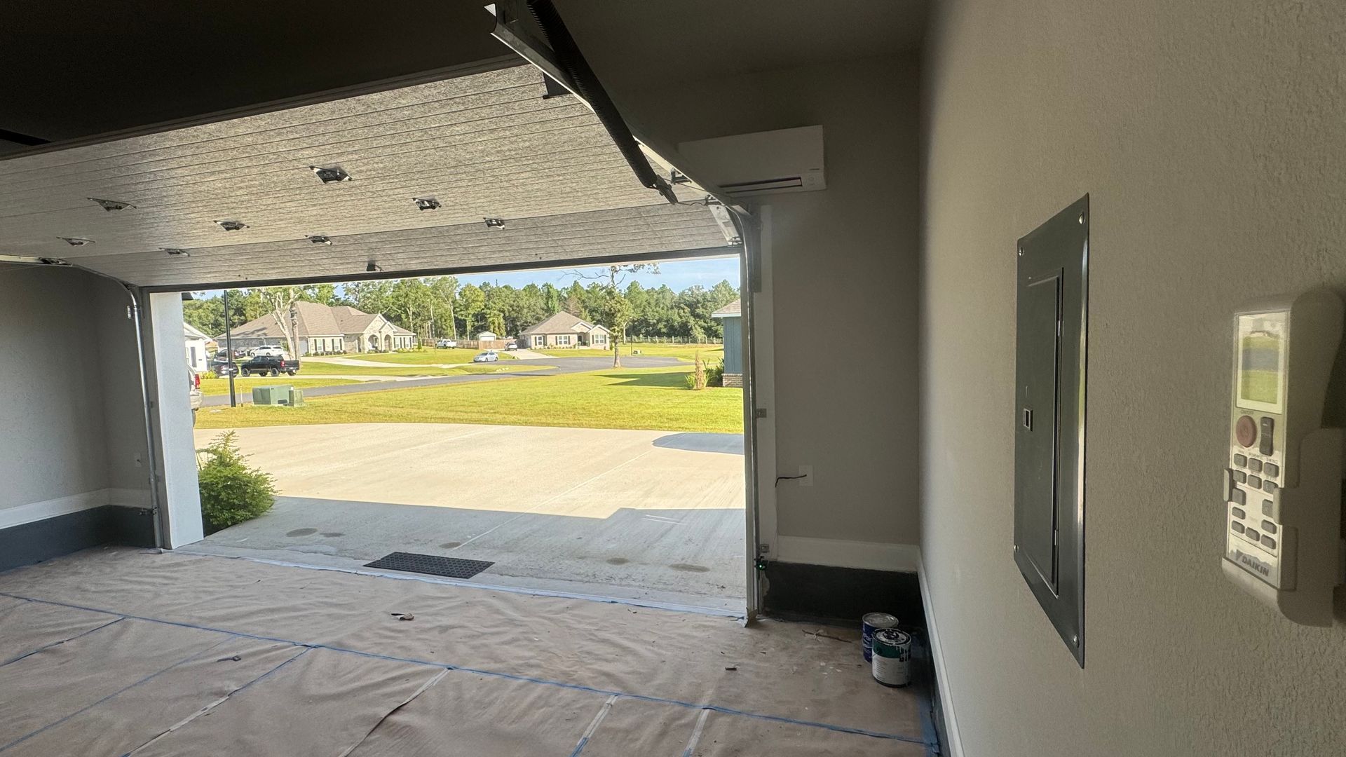 Open garage with view of neighborhood, panel box and thermostat on wall, paint can on ground.
