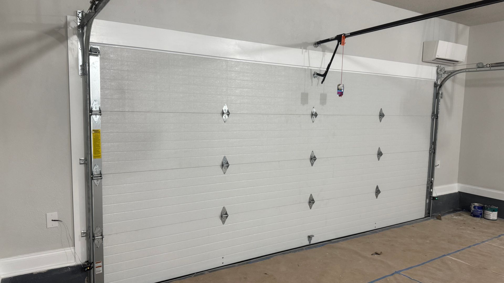 White garage door, closed, in a garage. Metal frame, gray walls, and a visible opener.