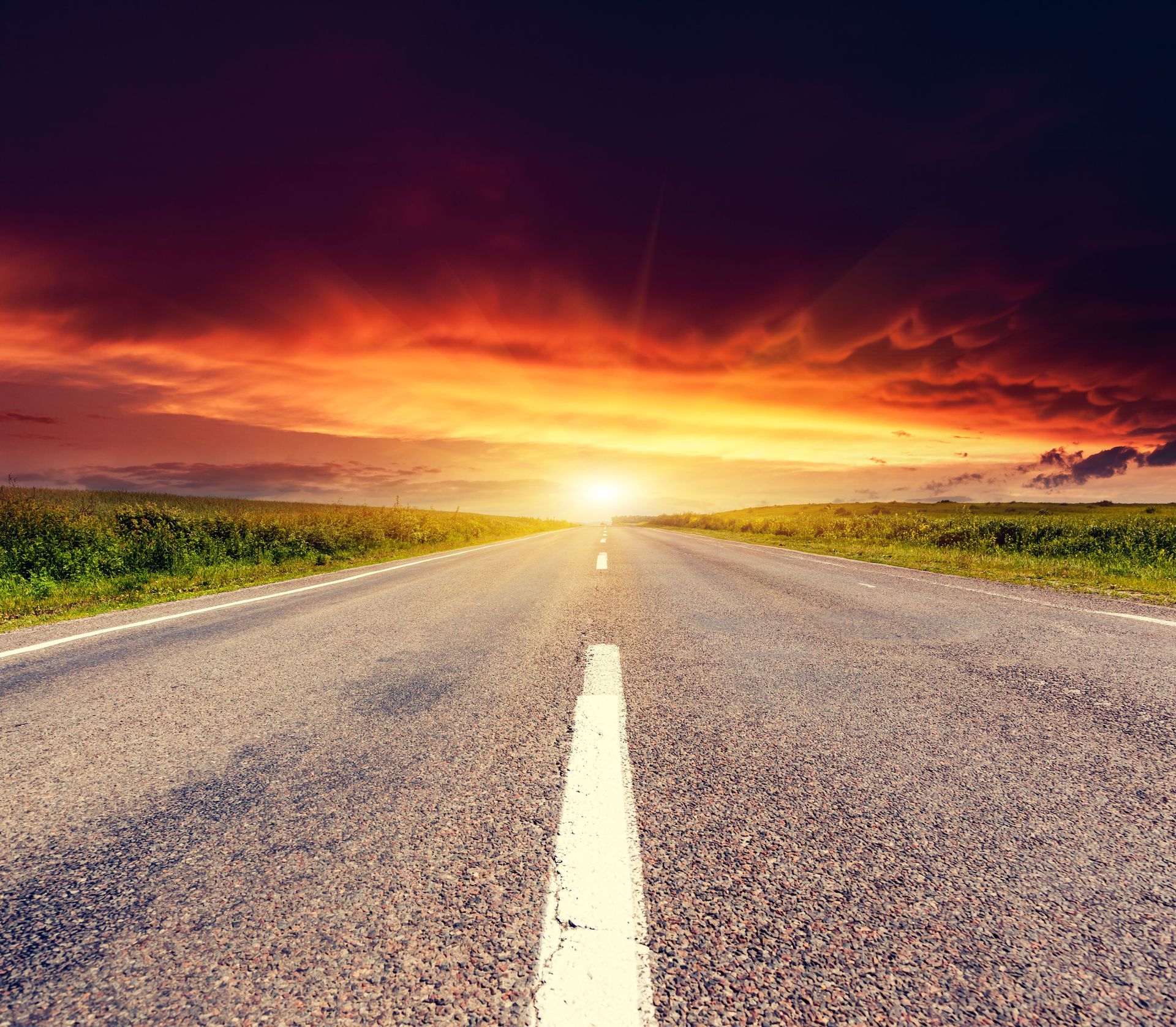 A long, empty asphalt road stretches toward a glowing sunset over a vast, grassy field under a dramatic, dark sky.