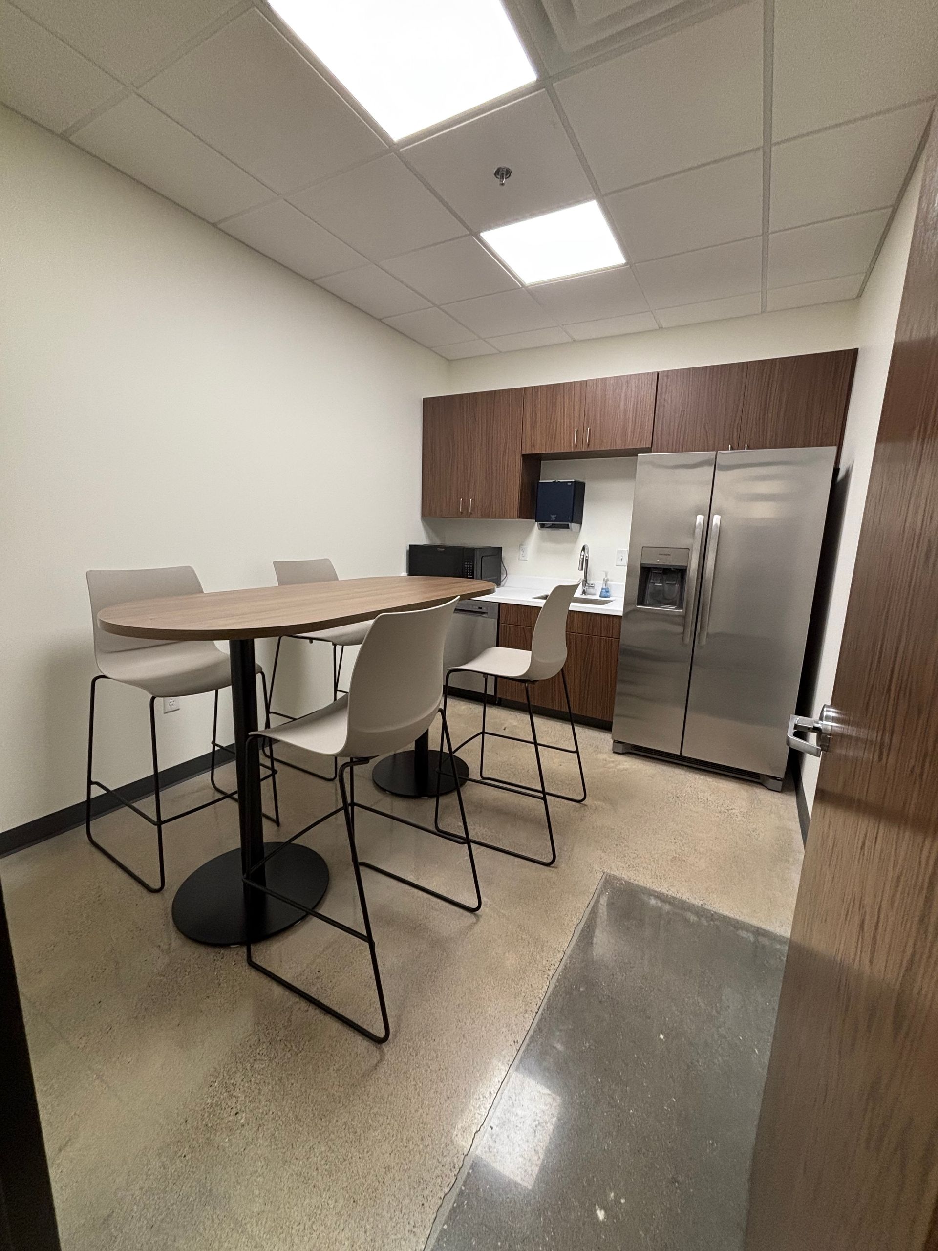 A small break room with a high table, four light-colored bar stools, brown cabinets, and a stainless steel refrigerator.