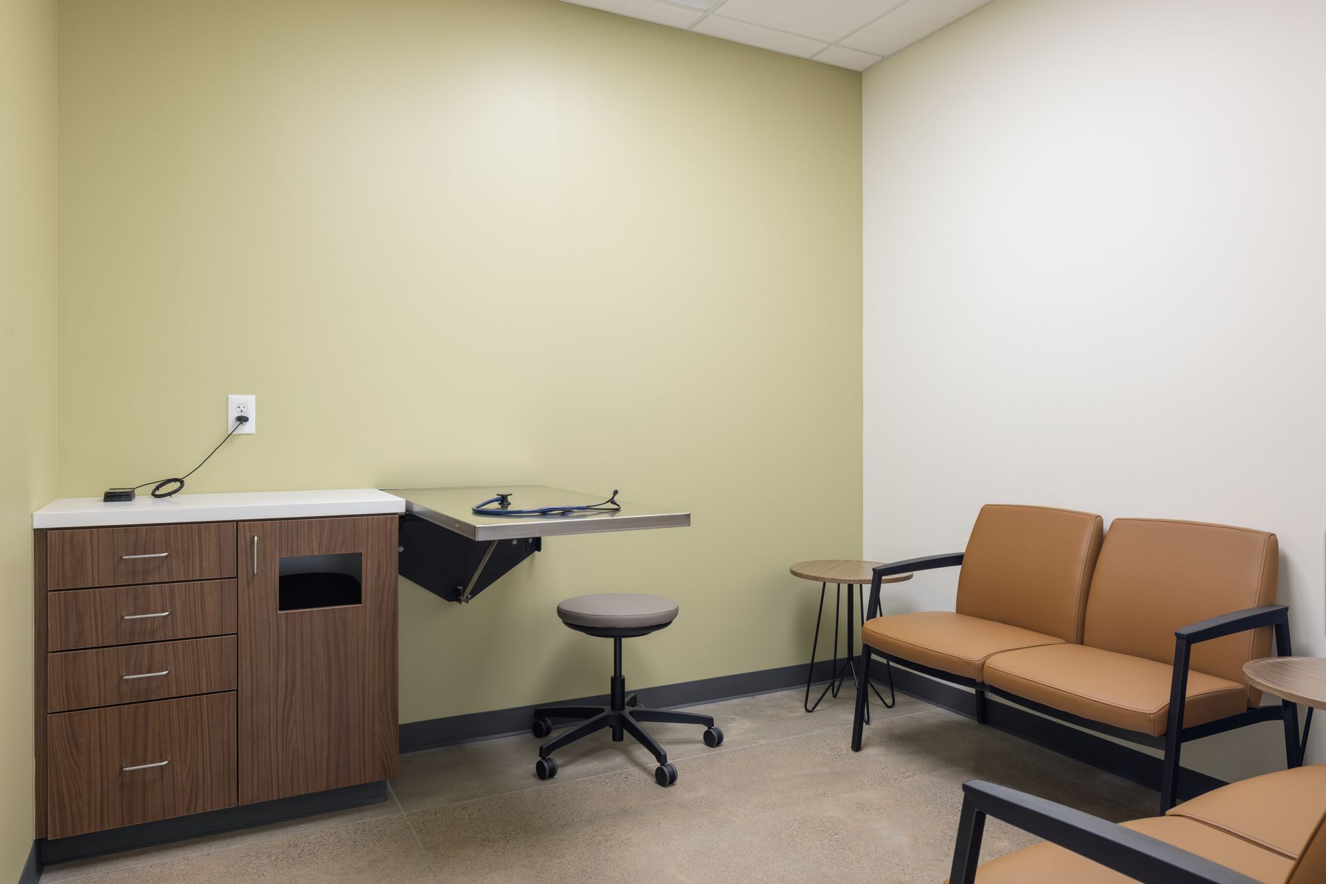 Examination room with tan walls, brown cabinets, steel table, and tan seating.