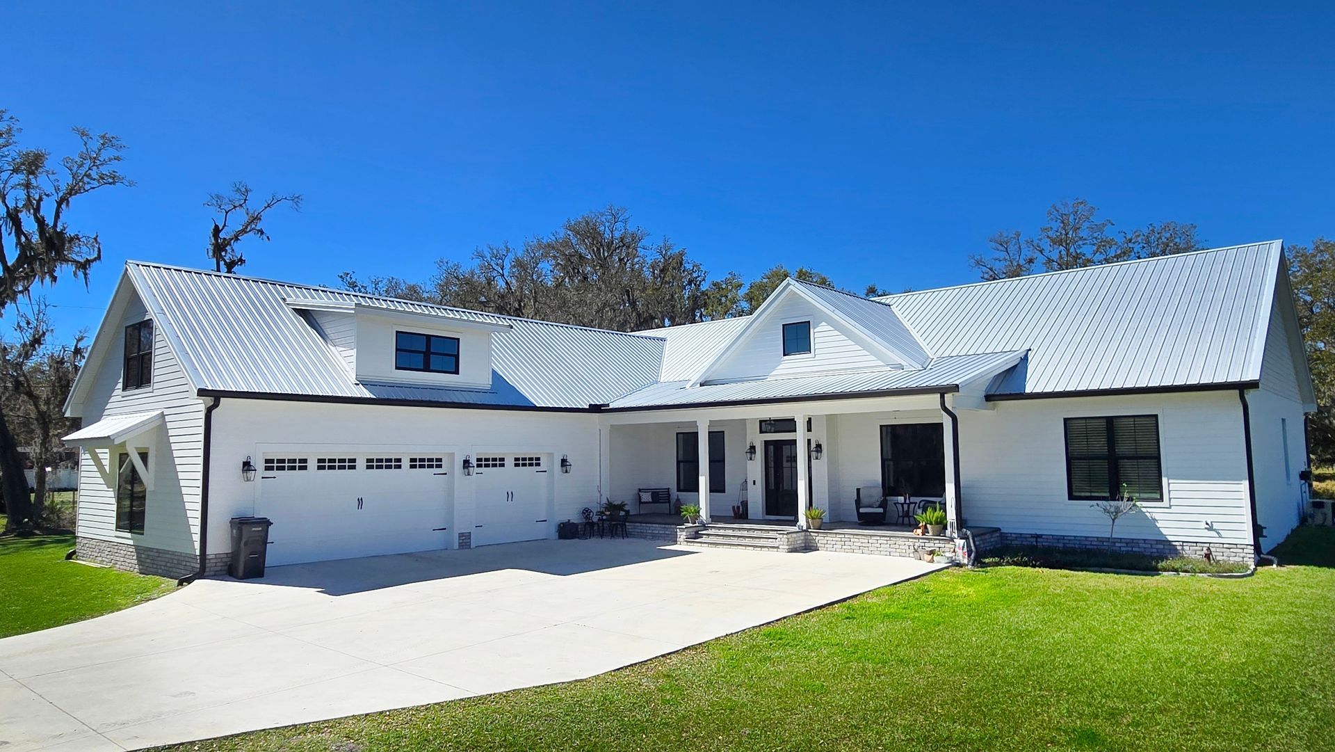 White farmhouse with metal roof, driveway, and green lawn under a blue sky.