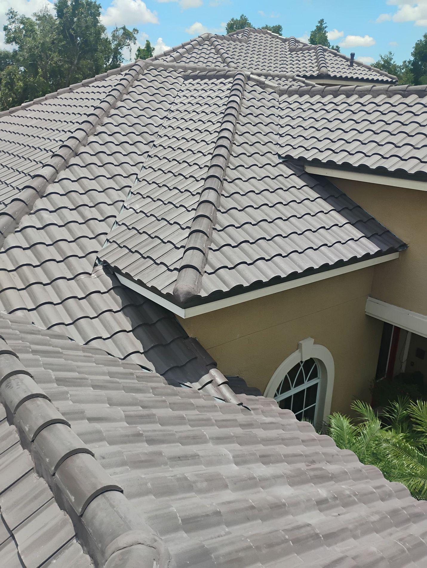 Gray tile roof on a beige house against a blue sky with trees.